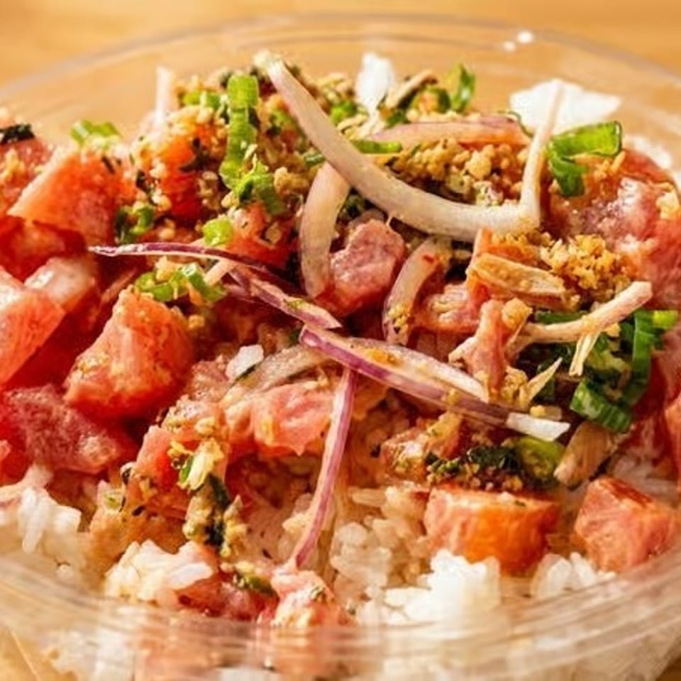 Close-up of a poke bowl with diced tuna, red onions, green onions, and rice in a clear container.