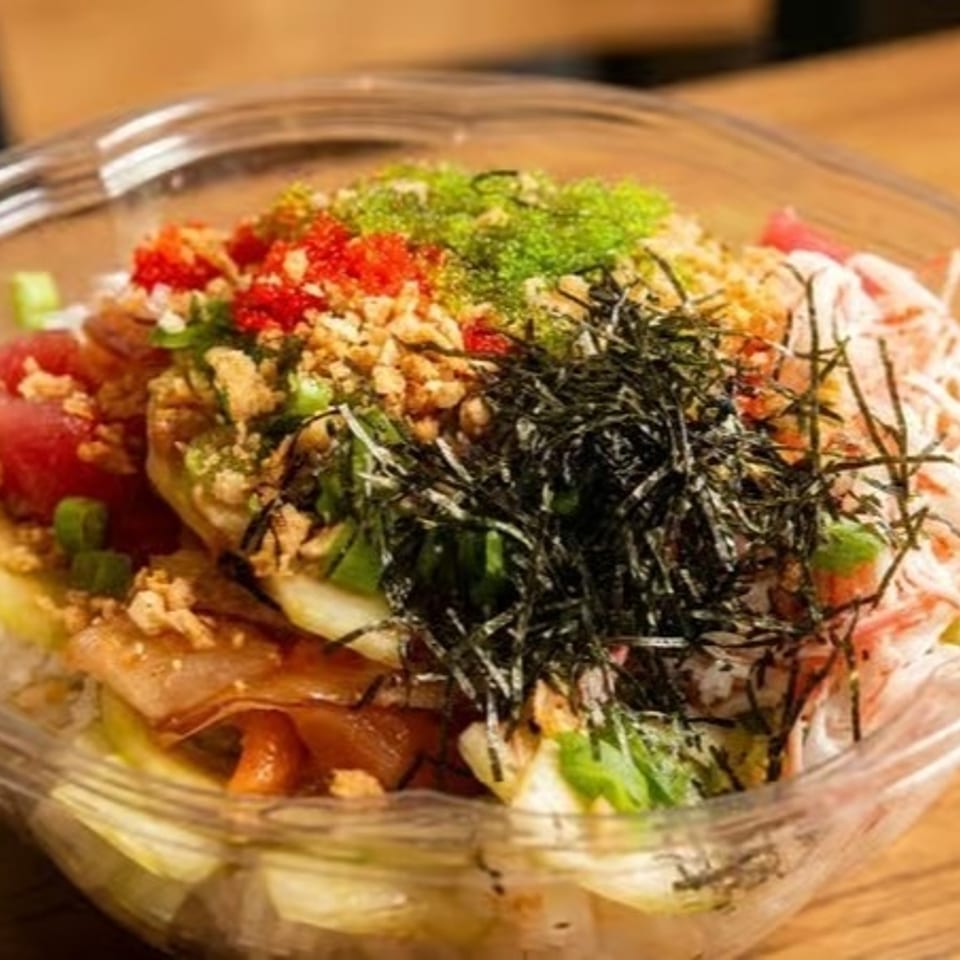 Colorful poke bowl with tuna, crab, seaweed, roe, and crunchy toppings in a clear plastic container.